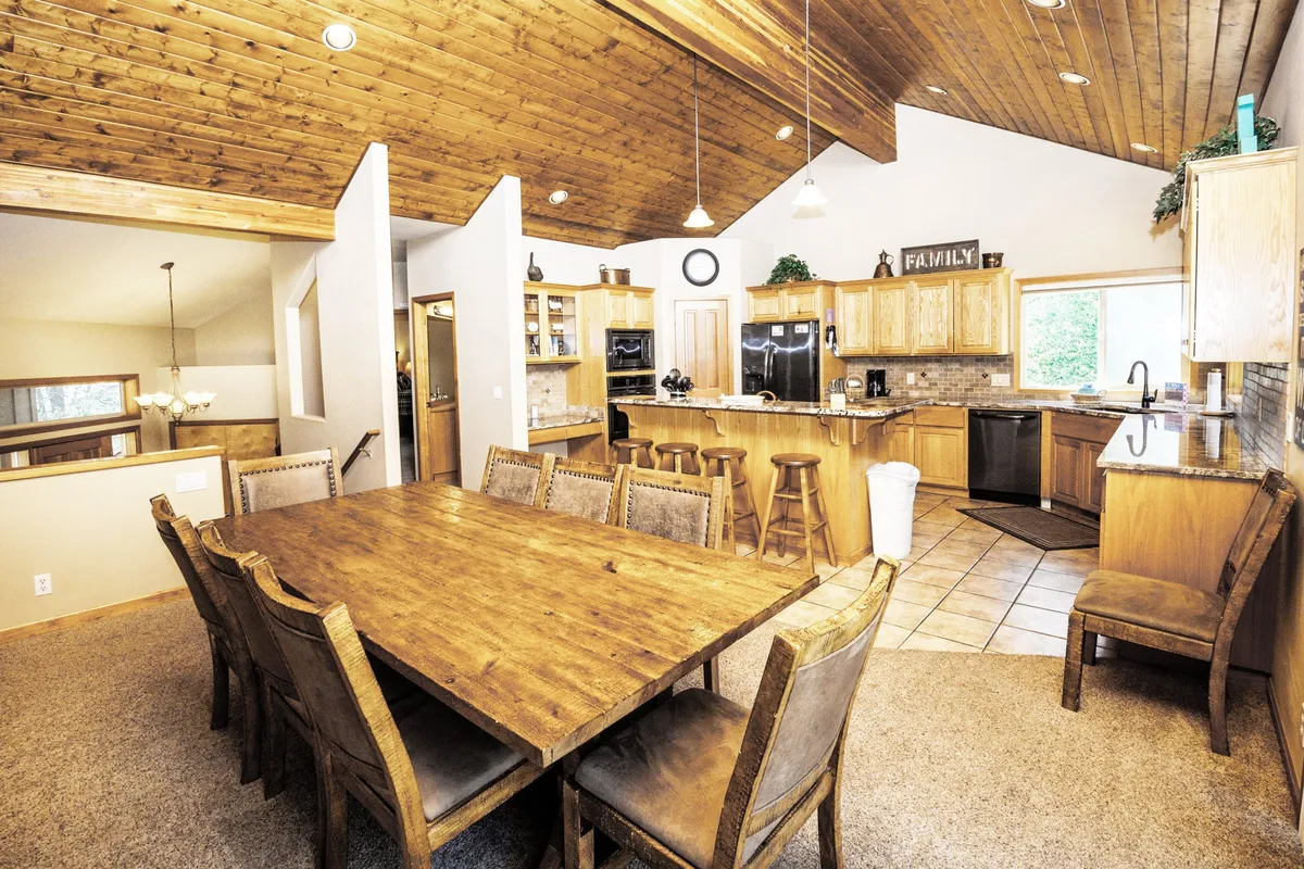 A large wooden dining table is set beneath a high wooden ceiling, surrounded by an open kitchen layout and bathed in natural light from generous windows. The setting radiates warmth and sets the stage for shared meals, celebrations, and casual conversations.