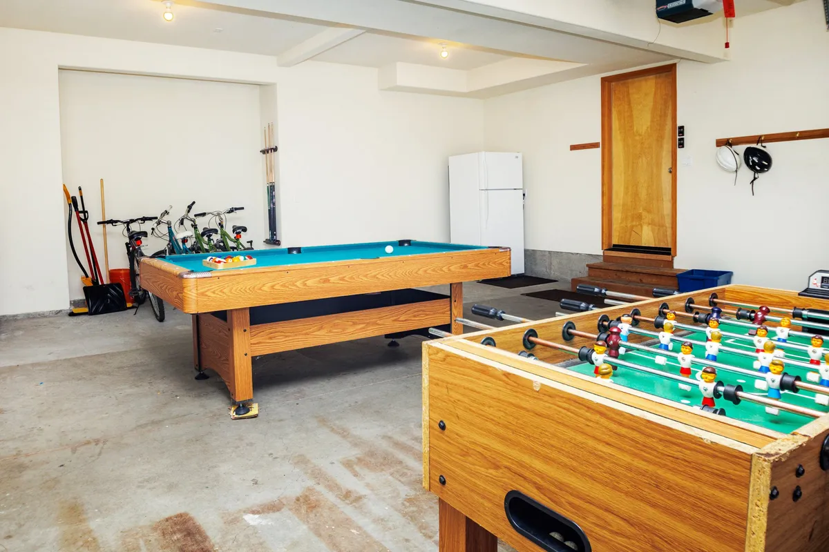 This image presents another lively view of the garage entertainment space, again highlighting a pool table and foosball table. A refrigerator and bicycles in the background add elements of convenience and versatility, showing that the garage is used for both recreation and practical storage. The mood is relaxed and social, with the space inviting for both gameplay and celebration.