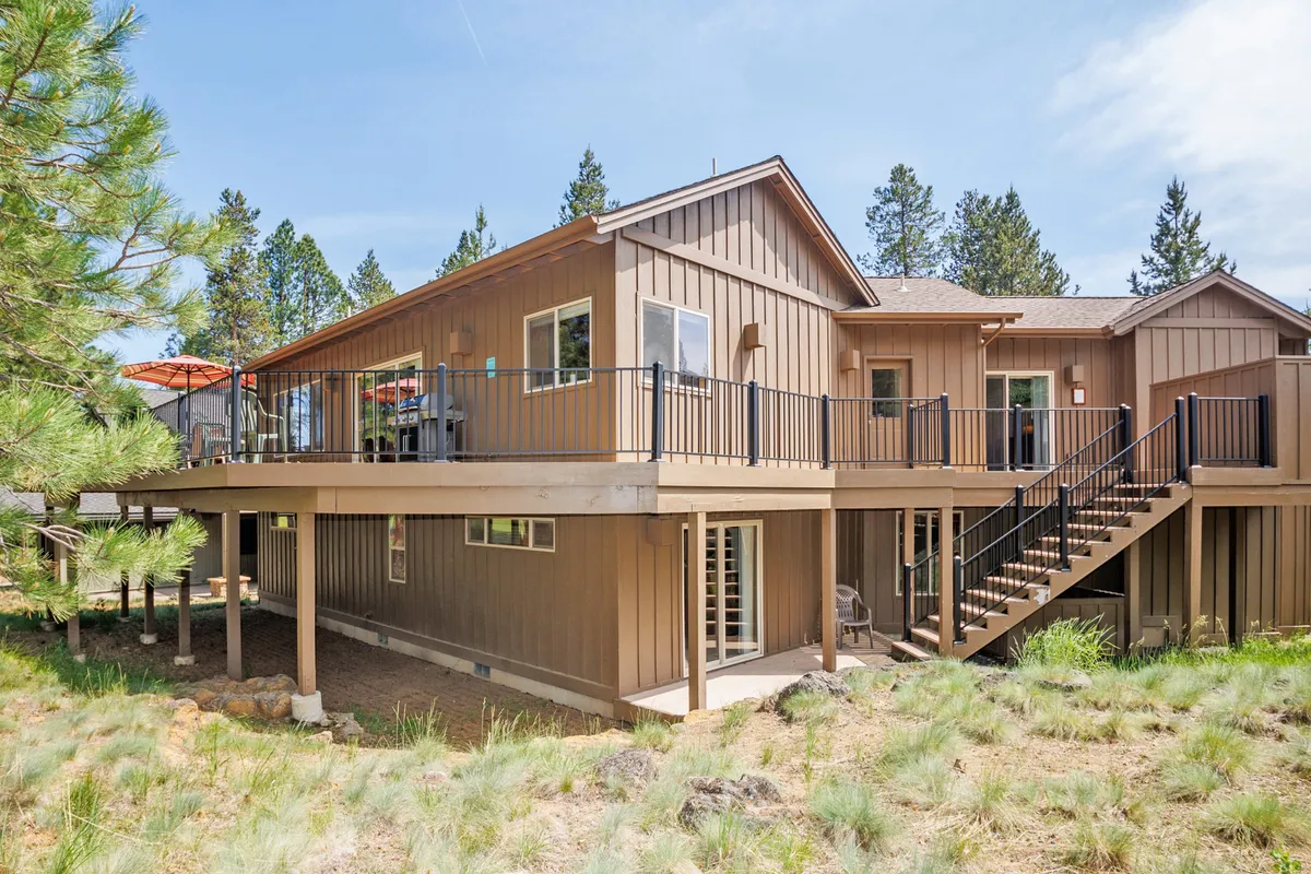 A broad exterior view of the home's deck area, bordered by a staircase and surrounded by lush greenery. The deck is spacious, offering ample room for outdoor dining and relaxation, with the natural landscape creating a tranquil backdrop ideal for enjoying the outdoors.