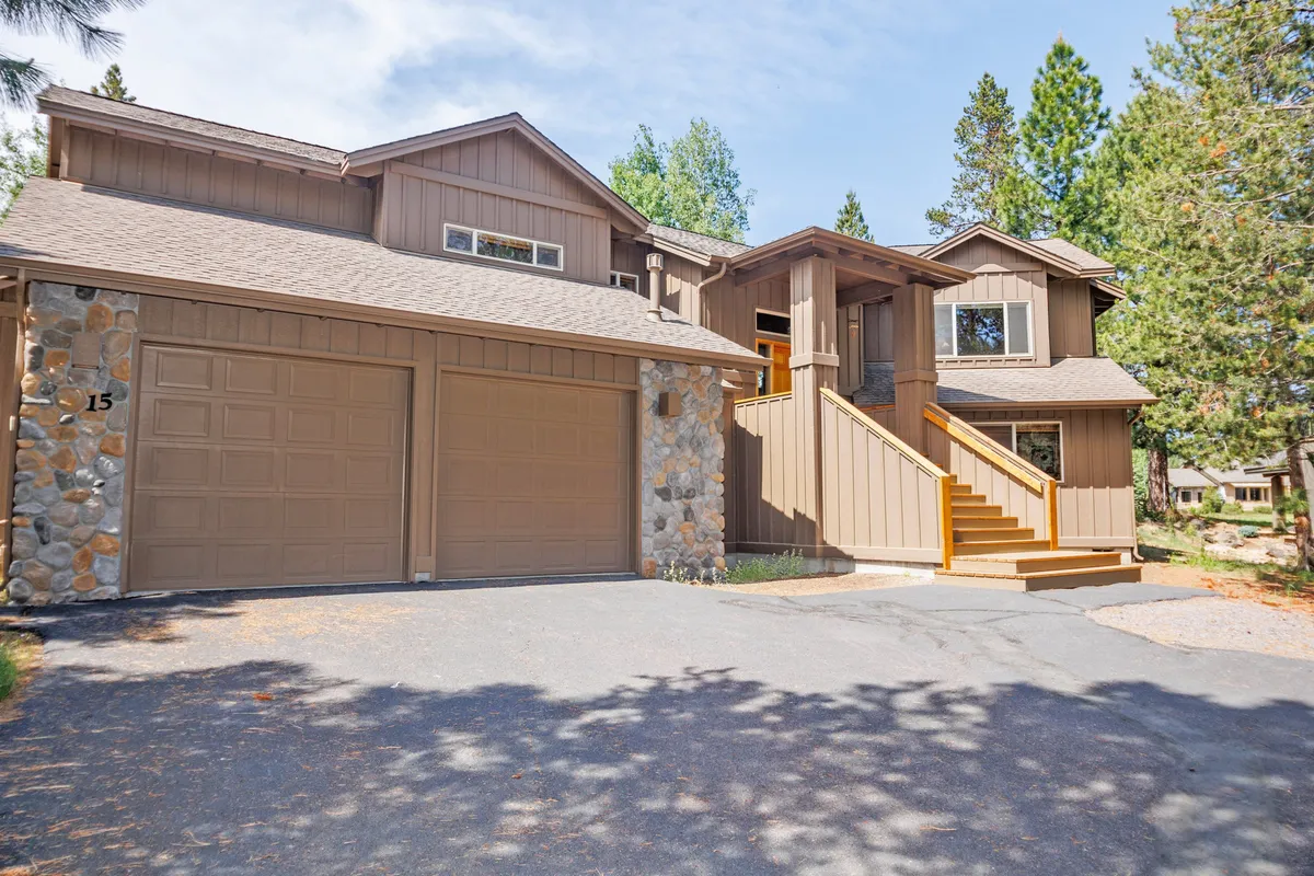 This image depicts the exterior of Tokatee 15, focusing on wooden steps leading to the entrance, stone accents on the garage, and tall trees in the background. The scene is serene and inviting, suggesting a welcoming arrival and a connection with the natural surroundings.