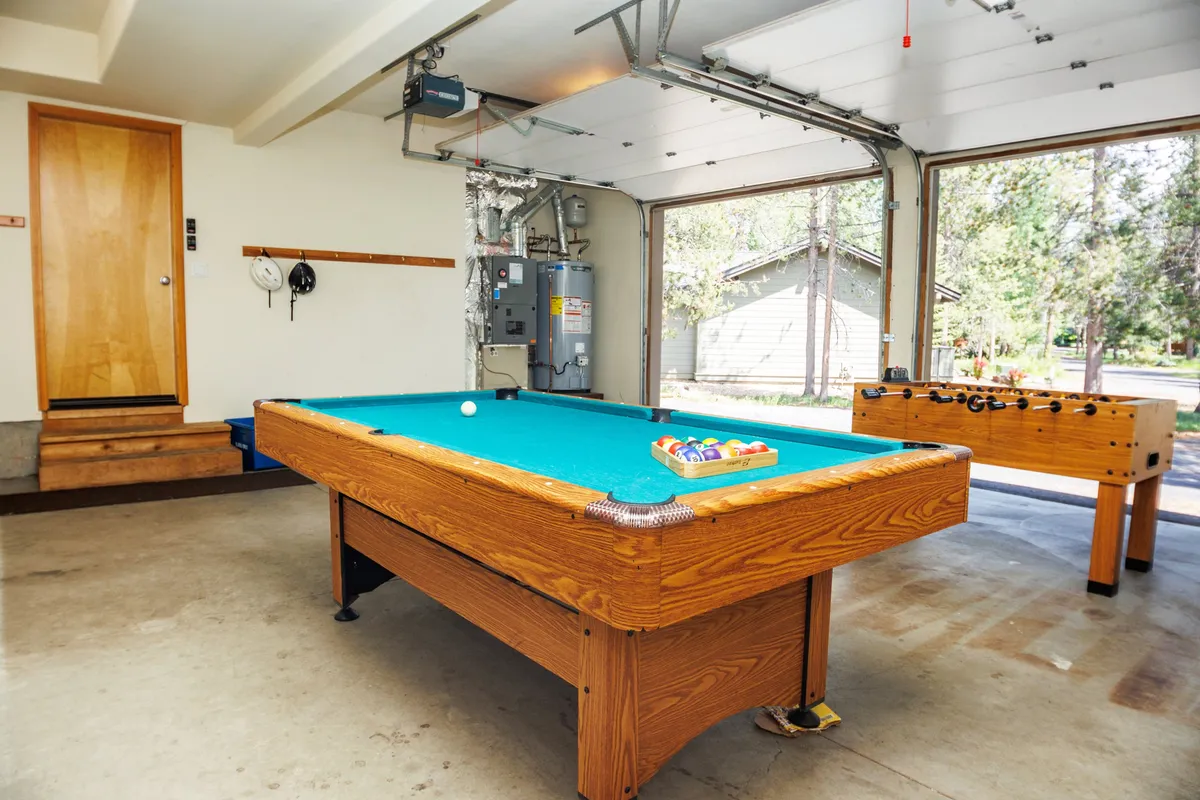 This image showcases a vibrant garage entertainment area featuring both a pool table and a foosball table. The garage door and wooden door add to the oversized, flexible nature of the space, while inviting lighting emphasizes the playful and relaxed atmosphere. The setup is clearly designed for group games, family bonding, and social gatherings.