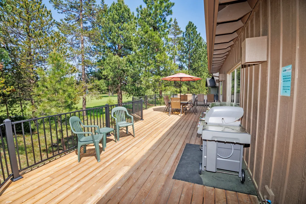 This image shows a spacious wooden deck furnished with green chairs, a barbecue grill, and a dining table with an umbrella. The setting is relaxed and social, surrounded by trees that evoke the area's natural beauty.