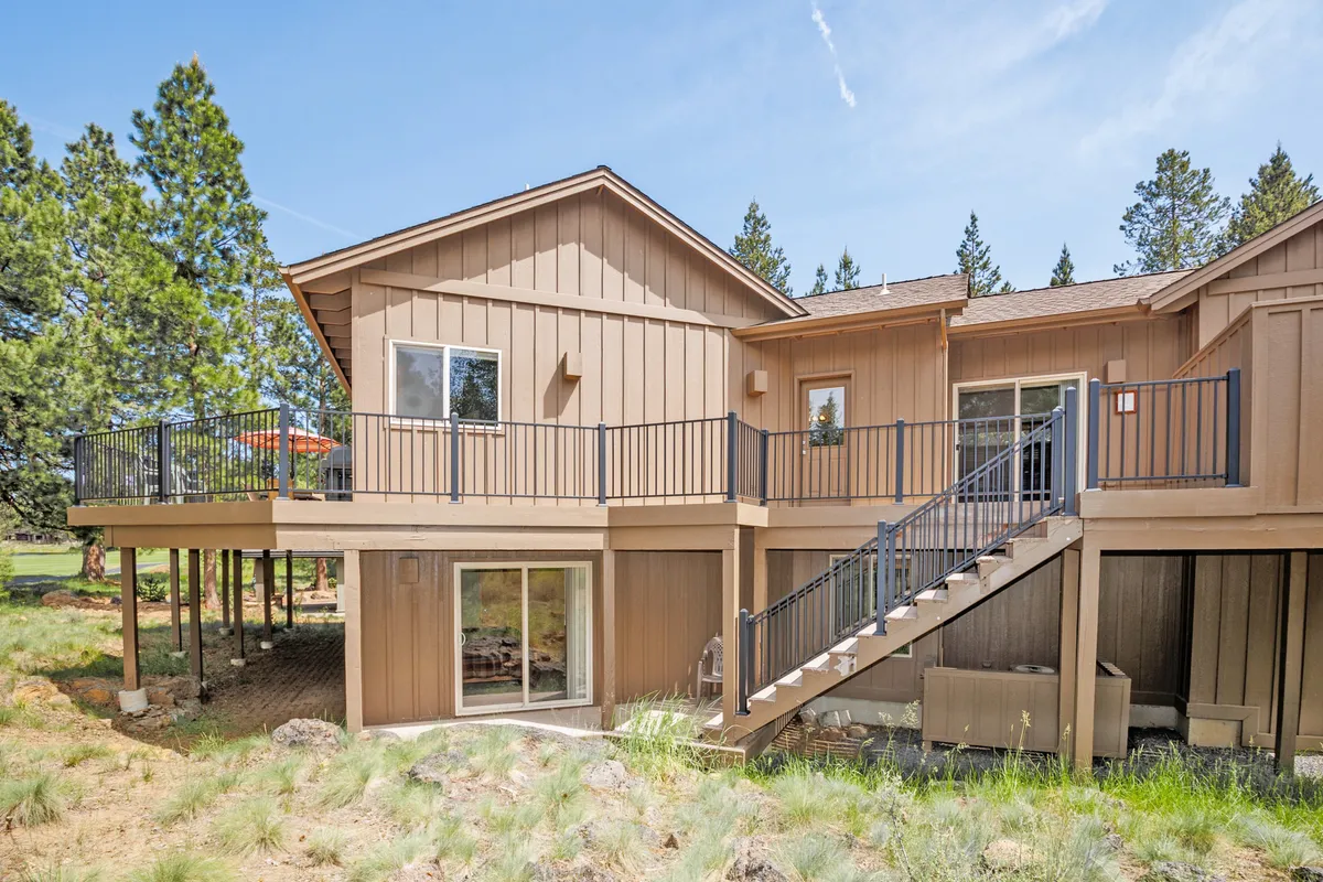 This exterior image showcases the home's spacious deck area, complete with a staircase leading down, large windows framing the house, and lush surrounding trees. The scene hints at a tranquil spot for morning coffee, group gatherings, or unwinding after a day of Sunriver adventures.