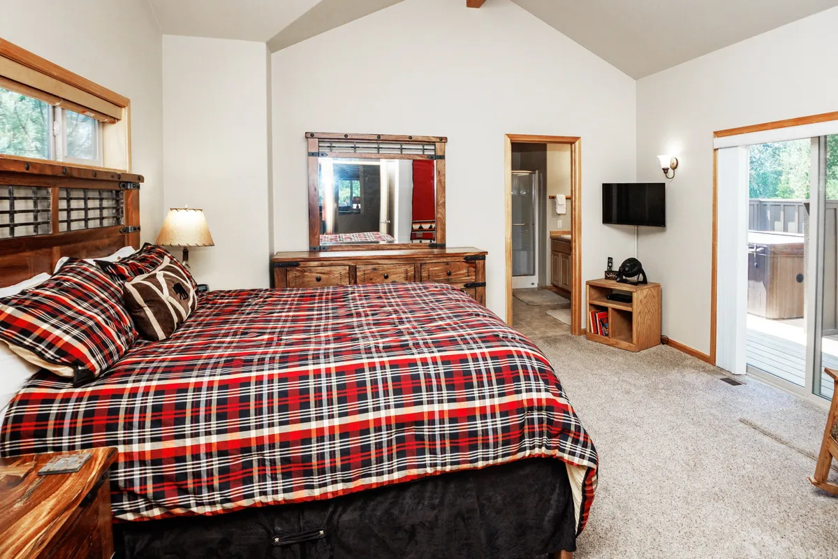 This bedroom features a plaid bedspread, wooden furniture, a large mirror, and a sliding glass door that lets in abundant natural light. The cozy decor and view through the door create a peaceful, restful atmosphere ideal for relaxation and privacy.
