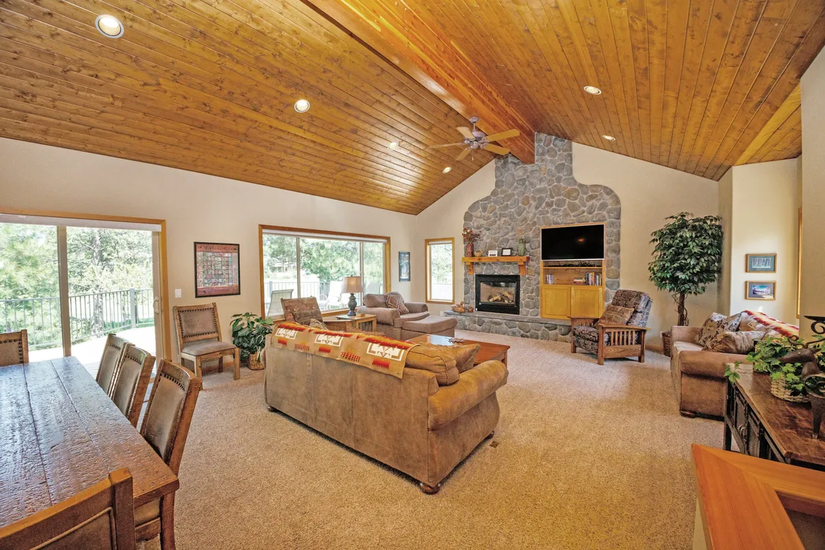 This image showcases a spacious living room with a stunning stone fireplace as the focal point. Large picture windows fill the room with natural light and provide beautiful views of the outdoors, while the high wooden ceiling adds to the sense of openness. Comfortable seating is arranged to invite relaxation and togetherness, making it ideal for family gatherings.