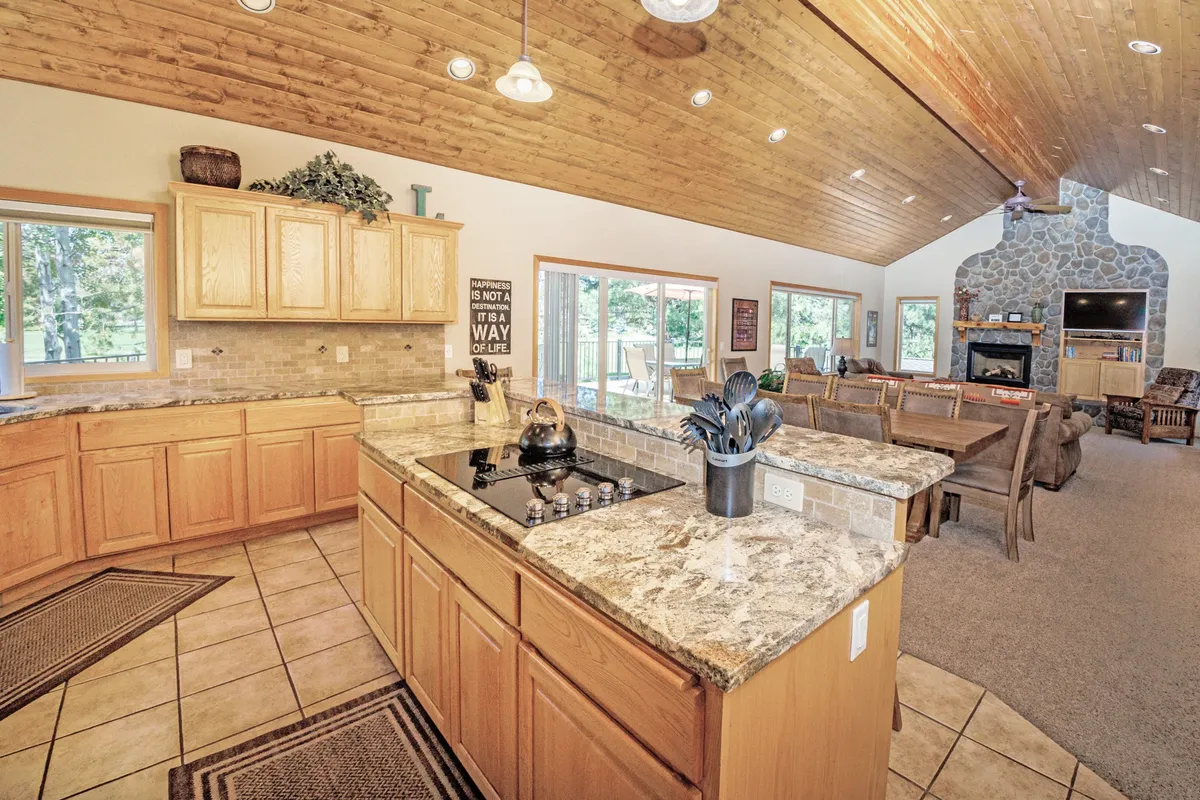 This photograph captures the open-concept living area, where a kitchen island with a stove flows seamlessly into a dining area and living space anchored by a stone fireplace. Large windows invite natural light, and the layout encourages conversation and connection among guests.